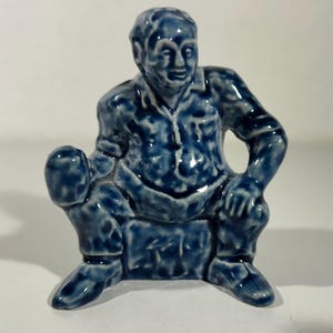 Könnte beinhalten: Eine blaue Keramikfigur, die einen sitzenden Mann darstellt. Die Figur trägt ein Hemd und eine Hose, die Arme ruhen auf den Beinen. Die Figur hat eine glänzende Oberfläche und ist in einem gesprenkelten Blau glasiert.