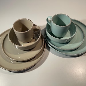 Könnte beinhalten: Ein Set aus zwei Geschirrkollektionen, die jeweils aus einer Tasse, einer Schüssel und einem Teller bestehen. Ein Set ist beige, das andere hellblau. Beide Sets haben eine glatte Oberfläche mit gesprenkelten, strukturierten Rändern.