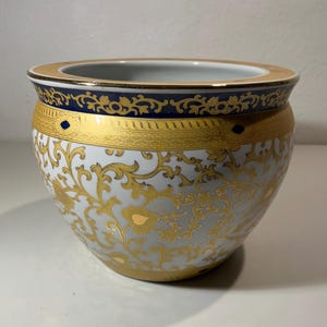 Könnte beinhalten: Ein weißer Keramikübertopf mit einem komplizierten goldenen Rankenmuster auf dem Körper. Der obere Rand ist mit einem breiten marineblauen Band verziert, das durch goldene Filigranarbeit und kleine dunkelblaue Rauten akzentuiert wird.