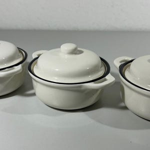Könnte beinhalten: Drei kleine, weiße Keramik-Auflaufformen mit Deckel und Griffen. Jedes Gericht hat eine schwarze Umrandung um den Rand und den Deckel. Die Auflaufformen sind in einer Reihe angeordnet, mit den Deckeln oben.