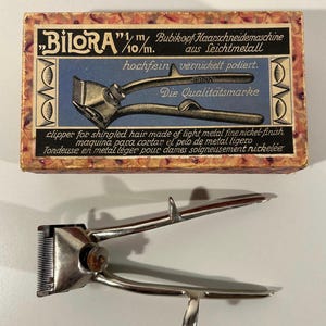 Könnte beinhalten: Vintage Haarschneidemaschine aus Metall mit Nickel-Finish, neben der Originalverpackung. Die Box zeigt Text in mehreren Sprachen, darunter "BILORA" und "clipper for shingled hair". Die Haarschneidemaschine ist zum Haareschneiden bestimmt.