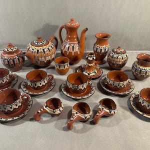 Könnte beinhalten: Ein Set aus Terrakotta-Keramikgeschirr mit brauner Glasur und dekorativen Tropfenmustern in Weiß, Blau und Schwarz. Das Set umfasst eine Teekanne, einen hohen Krug, eine Zuckerdose, ein Milchkännchen, Tassen mit Untertassen und kleine Schalen.