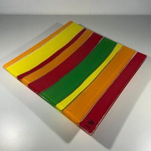 Könnte beinhalten: Ein quadratischer Glasteller mit einem lebendigen Streifendesign. Der Teller hat abwechselnde Bänder in Gelb, Orange, Rot und Grün. Der Teller hat eine glänzende Oberfläche und ein kleines Etikett in der Ecke.