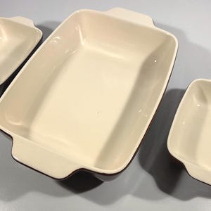 Könnte beinhalten: Set aus drei cremefarbenen Keramik-Backformen. Das Set enthält eine große rechteckige Form und zwei kleinere quadratische und rechteckige Formen. Jede Form hat einen dunkelbraunen Rand und Griffe.