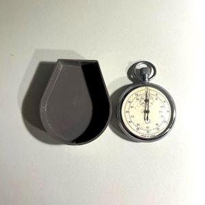 Könnte beinhalten: Eine Vintage-Stoppuhr mit silberfarbenem Gehäuse und cremefarbenem Zifferblatt. Das Zifferblatt hat schwarze Ziffern und Markierungen, mit einem Sekundenzeiger und einem kleineren Zifferblatt. Die Stoppuhr liegt neben einem grauen, tropfenförmigen Gehäuse.