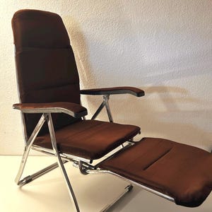 May include: A folding recliner chair featuring a shiny chrome metal frame and dark brown fabric upholstery. The chair includes a high backrest, two armrests with black accents, a seat, and an integrated footrest that extends outward.
