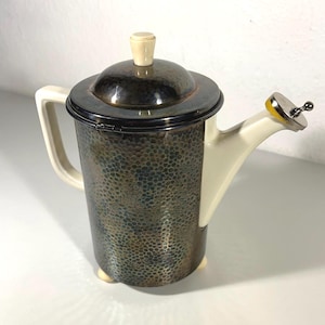 May include: A vintage silver-toned metal teapot with a textured surface and a cream-colored handle and spout. The lid has a cream-colored knob. The teapot has a unique design and is likely from the mid-century era.