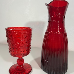 Könnte beinhalten: Ein roter Glasbecher und eine passende Karaffe. Der Becher hat ein detailliertes Muster und einen runden Fuß. Die Karaffe hat ein geriffeltes Design und einen ausgestellten oberen Rand. Beide Artikel sind aus rotem Glas.