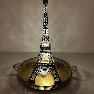 May include: A decorative Eiffel Tower figurine with internal lights, set on a gold-colored metal tray. The tower is black with intricate lattice detailing, and the lights create a warm glow. The tray has ornate handles.