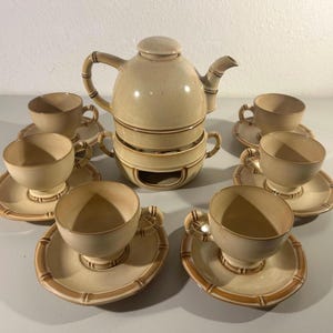 Può includere: Un servizio da tè in ceramica beige con una teiera, sei tazze e piattini. La teiera ha un manico e un beccuccio in stile bambù. Le tazze e i piattini hanno un bordo in stile bambù. Il set è disposto su una superficie chiara.