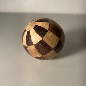 Könnte beinhalten: Ein Holzpuzzle-Ball mit einem geometrischen Muster aus abwechselnd hell- und dunkelbraunen Quadraten. Der Ball ist kugelförmig und scheint aus ineinandergreifenden Teilen zu bestehen. Das Design ist symmetrisch und optisch ansprechend. Durchmesser: ca. 7 cm.