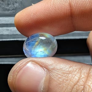 Puede incluir: Un cabujón de piedra lunar de forma ovalada y lisa que presenta un aspecto translúcido con una distintiva adularescencia o brillo azul. La piedra preciosa muestra inclusiones internas y una superficie pulida que refleja la luz.
