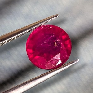 May include: A round, faceted, deep red ruby gemstone held by a pair of silver-toned metal tweezers. The gemstone displays a vibrant crimson hue with visible internal inclusions and a polished surface that reflects light across its circular cut.