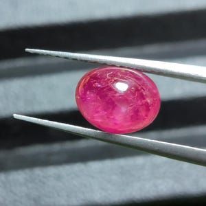 Może przedstawiać: Żywy, owalny, półprzezroczysty różowo-czerwony kamień szlachetny trzymany przez srebrną metalową pęsetę. Kamień ma gładką, polerowaną powierzchnię typu kaboszon z widocznymi wewnętrznymi inkluzjami oraz jasnym punktem świetlnym na górze.