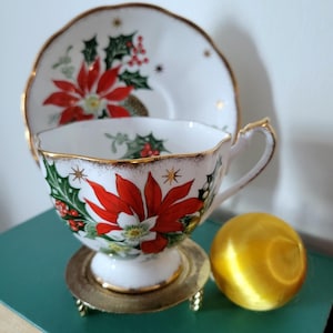 May include: A white teacup and saucer set with gold edging, decorated with red poinsettias and holly. The teacup is on a small gold stand, with a gold sphere beside it. The set is likely for festive occasions.