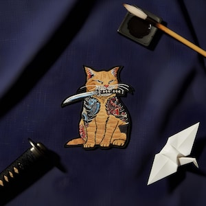 Peut inclure: Écusson brodé représentant un chat roux souriant avec des tatouages, tenant un katana dans sa bouche. Le chat est entouré d'un pinceau de calligraphie, d'encre, d'un katana et d'une grue en papier, sur fond bleu marine.