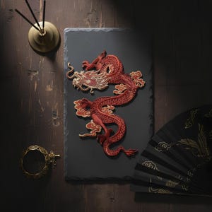 May include: A detailed, red embroidered dragon patch with gold accents, displayed on a dark gray slate. The dragon is the focal point, with a black fan and gold incense holder in the background. The dragon is a symbol of power and good fortune.