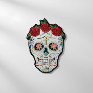 Könnte beinhalten: Gestickter Totenkopf-Aufnäher mit floralem Design. Der weiße Totenkopf hat rote Rosen, rosa und grüne Blumen und ein goldenes Kreuz. Der Aufnäher hat einen schwarzen Rand und ist wahrscheinlich für Kleidung oder Accessoires.