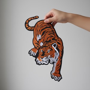 Può includere: Toppa ricamata a forma di tigre in arancione, nero e bianco. La tigre è raffigurata in posizione accovacciata con strisce e tratti del viso dettagliati. La toppa è tenuta su uno sfondo neutro.