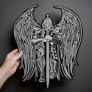 Può includere: Grande toppa ricamata in bianco e nero raffigurante un angelo guerriero in ginocchio con una spada. L'angelo ha grandi ali dettagliate e indossa un'armatura. La toppa è tenuta da una mano su uno sfondo grigio.