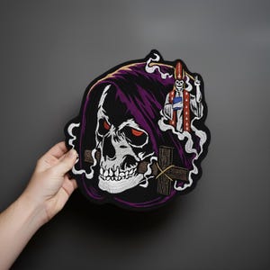 Può includere: Toppa ricamata raffigurante un teschio con occhi rossi, un cappuccio viola e una figura in una veste rossa e bianca. Il disegno comprende una croce e accenti bianchi a forma di fumo su sfondo nero. La toppa è tenuta in mano.