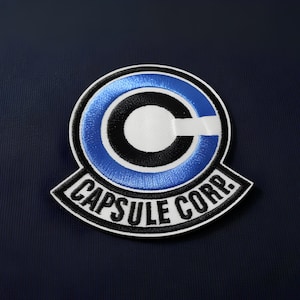 May include: Embroidered patch with the Capsule Corp logo from Dragon Ball. The patch features a white, black, and blue design. The text "CAPSULE CORP." is displayed in white on a black banner.