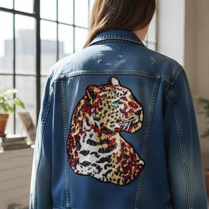 Può includere: Giacca di jeans blu medio con una grande toppa di leopardo con paillettes sul retro. La toppa mostra la testa di un leopardo con paillettes rosse, dorate e bianche e occhi verdi. La giacca ha classici dettagli in denim.