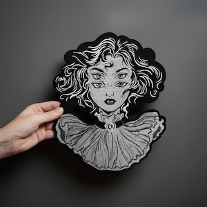 May include: Embroidered patch depicting a woman with four eyes, curly hair, and a ruffled collar, in black and white. The detailed design is held against a grey background. The patch is a unique, gothic-inspired accessory.