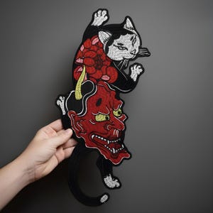 Peut inclure: Écusson brodé représentant un chat noir avec des détails blancs sur un masque de démon rouge. Le chat a un motif floral sur le dos. Le masque de démon a des yeux et des cornes jaunes. Le patch est un design unique.