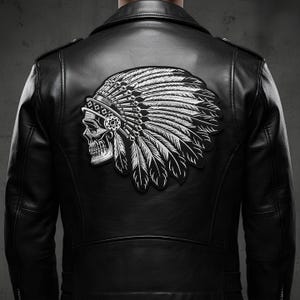 Może przedstawiać: Czarna skórzana kurtka z dużą naszywką przedstawiającą czaszkę w indiańskim nakryciu głowy. Naszywka jest biało-szara, ze szczegółowymi piórami i detalami czaszki. Kurtka ma klasyczny styl motocyklowy.