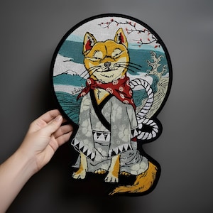 Puede incluir: Parche bordado con un perro Shiba Inu con un atuendo tradicional japonés, que incluye un pañuelo rojo y un kimono. El fondo representa un diseño de olas, montañas y flores de cerezo. El parche es redondo con un borde negro.
