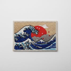 May include: Embroidered patch depicting a large ocean wave with a red sun. The wave is in shades of blue and white, set against a tan background with a white border. The patch is rectangular in shape.