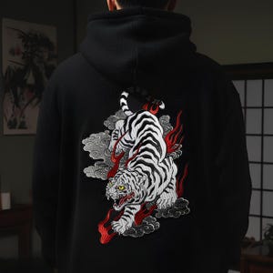 Puede incluir: Sudadera con capucha negra con un diseño bordado en la espalda. El diseño presenta un tigre blanco con rayas negras, llamas rojas y nubes grises. El tigre tiene ojos amarillos y está en una pose dinámica.