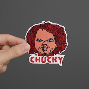 Può includere: Toppa ricamata raffigurante il volto di Chucky, con capelli rossi, occhi blu e un design cucito. La parola "CHUCKY" è visualizzata in rosso, delineata in bianco, sotto il volto.