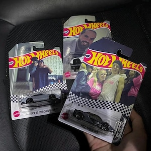 May include: Three Hot Wheels toy car packages. The packages feature images of people and the Hot Wheels logo. The cars are gray and black. One package says "Porsche 911 Carrera T" and another says "McLaren W1".