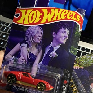 May include: Hot Wheels die-cast car featuring a red '71 Lamborghini Miura SV with gold wheels. The packaging includes a photo of a smiling couple. The name "ALEX" is printed on the side, and the date "03.09.24" is visible.