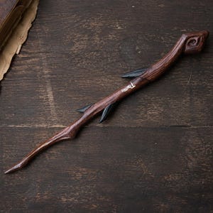 Puede incluir: Una varita de madera de color marrón oscuro con un mango tallado en espiral, una punta cónica y tres pequeñas protuberancias negras en forma de hoja a lo largo del cuerpo. El nombre Thomas está grabado en letras blancas en el lateral.