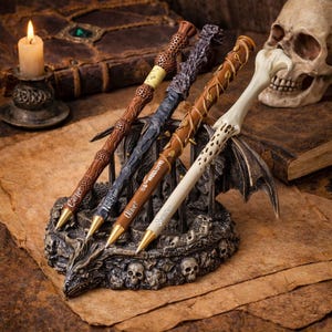 May include: Four decorative pens with intricate designs, displayed in a skull-themed holder. The pens feature various colors and textures, including brown, gray, and white, with gold-colored tips. A lit candle and a skull are in the background, creating a fantasy theme.