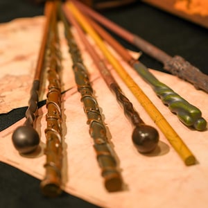 Può includere: Una collezione di bacchette artigianali in vari colori e design. Le bacchette sono fatte di legno e presentano dettagli intricati, tra cui manici scolpiti e accenti decorativi. Le bacchette sono disposte su una superficie simile a pergamena.