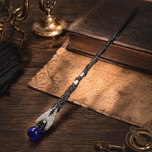 May include: A black wand featuring a textured, vine-like handle with a silver-toned metal accent at the base. The handle is adorned with a small white pearl and terminates in a vibrant, spherical blue glass orb held by silver-toned metal wings.