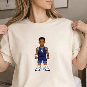 Pode incluir: Uma camiseta creme com uma ilustração de desenho animado de um jogador de basquete em uma camisa azul com o número 5. O jogador está usando shorts azuis, tênis brancos e pulseiras. O design é centralizado na camisa.