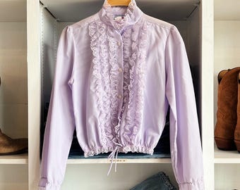 May include: A light purple long-sleeve blouse featuring a high collar, a vertical front placket with delicate white lace ruffles, a cinched elasticized waist with a drawstring tie, and matching lace trim on the cuffs and collar edges.