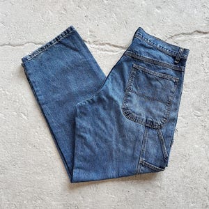 May include: A pair of blue denim carpenter pants. The jeans have a classic five-pocket design, with a hammer loop and a utility pocket on the right leg. The pants are folded and laid flat on a light gray surface.