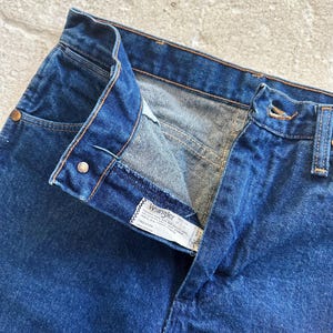 Pu&ograve; includere: Un paio di jeans blu denim con una cintura in pelle marrone e un'etichetta Wrangler. I jeans hanno una chiusura con bottoni e un solo bottone in vita.