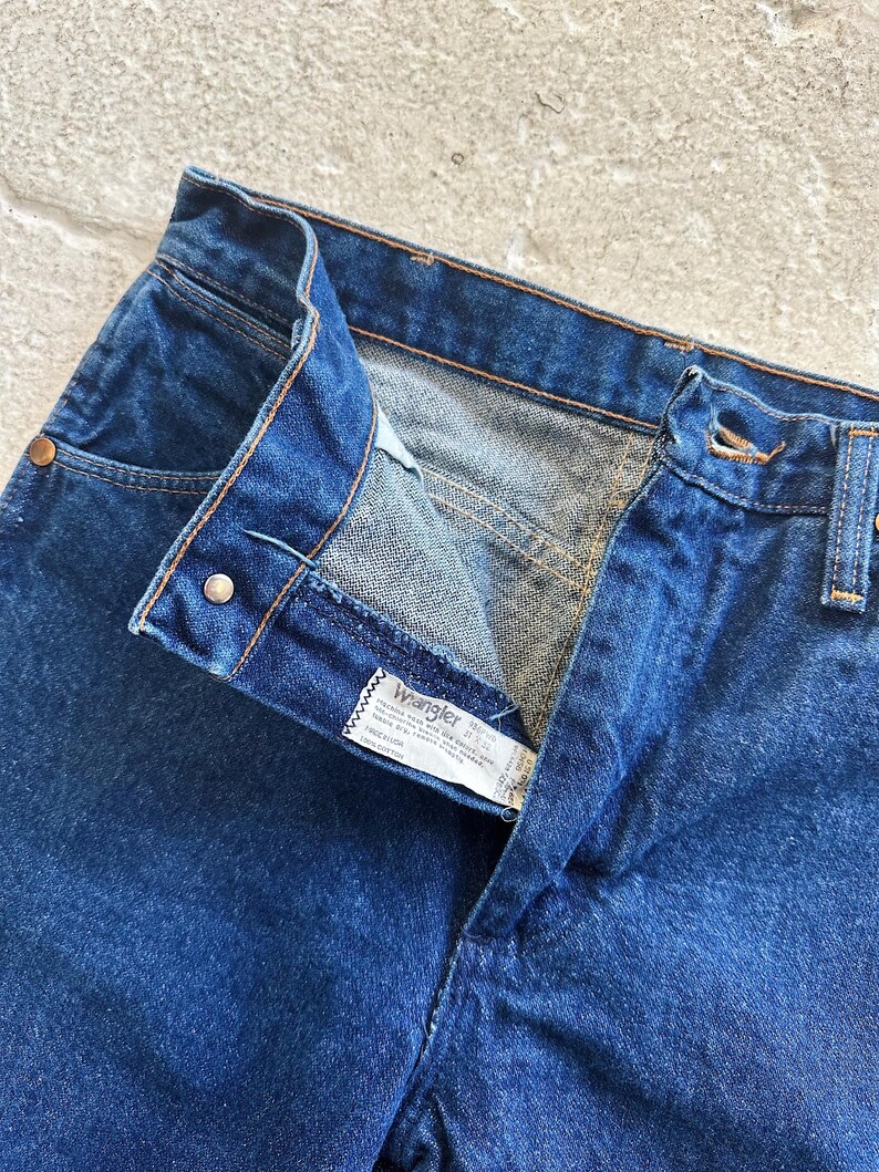 Pu&ograve; includere: Un paio di jeans blu denim con una cintura in pelle marrone e un'etichetta Wrangler. I jeans hanno una chiusura con bottoni e un solo bottone in vita.