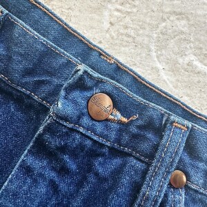 Pu&ograve; includere: Primo piano della cintura di un paio di jeans blu. La cintura ha un bottone color rame e un'asola color rame. I jeans sono realizzati in denim morbido e confortevole.