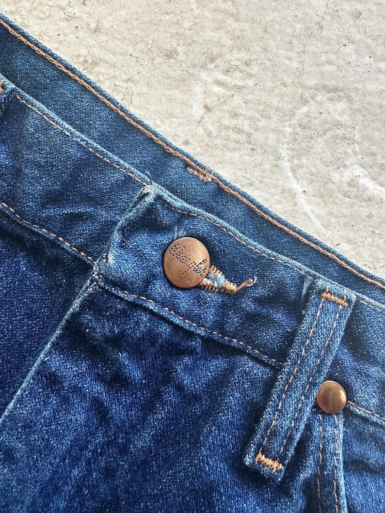 Pu&ograve; includere: Primo piano della cintura di un paio di jeans blu. La cintura ha un bottone color rame e un'asola color rame. I jeans sono realizzati in denim morbido e confortevole.