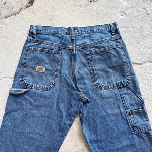 May include: Blue denim work pants with multiple pockets, including a side pocket and two back pockets. The pants have a classic, straight-leg fit and a small brand patch on the back pocket. The jeans are made of a durable denim material.