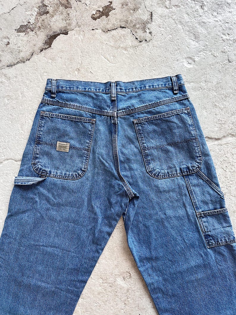 May include: Blue denim work pants with multiple pockets, including a side pocket and two back pockets. The pants have a classic, straight-leg fit and a small brand patch on the back pocket. The jeans are made of a durable denim material.