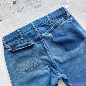 Puede incluir: Primer plano de la parte trasera de unos vaqueros azules. Los vaqueros tienen un aspecto descolorido con una etiqueta Wrangler en la cintura trasera. Los vaqueros tienen un bolsillo trasero y trabillas.